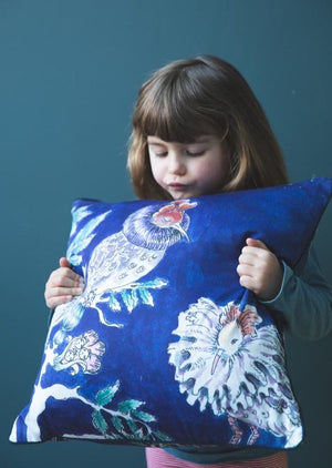 A little girl looking down and holding a square blue cushion featuring two hand illustrated ruff birds.