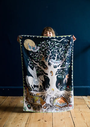 A smiling little girl holding a hand illustrated nighttime forest scene muslin blanket.
