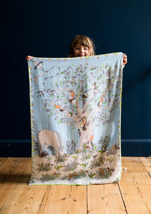 A smiling little girl holding a hand illustrated daytime forest scene muslin blanket.