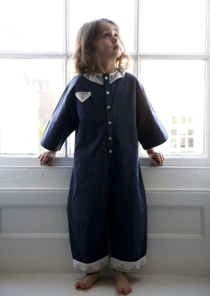 Little girl standing in front of a sash window wearing nighttime blue jumpsuit.