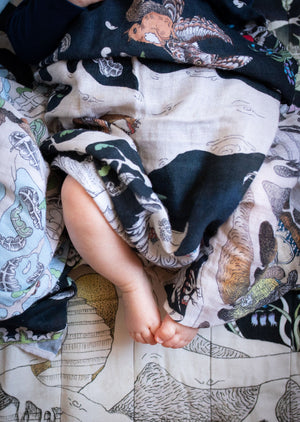 The bare feet of a newborn baby wrapped in a hand illustrated forest scene muslin blanket. 
