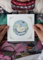Person holding a card with a swan illustration against a colorful fabric background