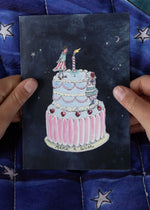 Person holding a card with a cake illustration against a blue quilted background.