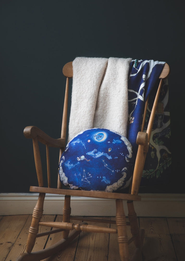 Round blue cushion with hand illustrated design featuring a nighttime sky design on a wooden chair.