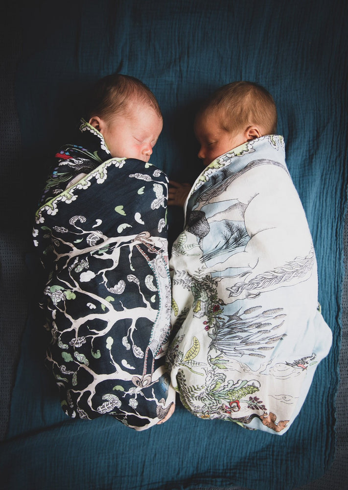 Twin newborn babies asleep, wrapped in hand illustrated forest scene muslin blankets. 