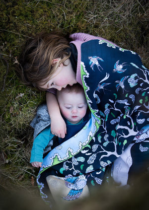 Little girl cuddling a baby under a reversible woodland scene organic muslin blanket.