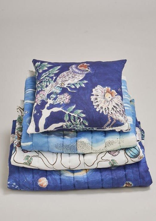 Organic cotton square blue cushion featuring two hand illustrated ruff birds on top of several quilted blankets.