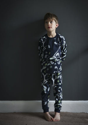 A little boy standing infront of a wall wearing woodland scene blue cotton pyjamas.